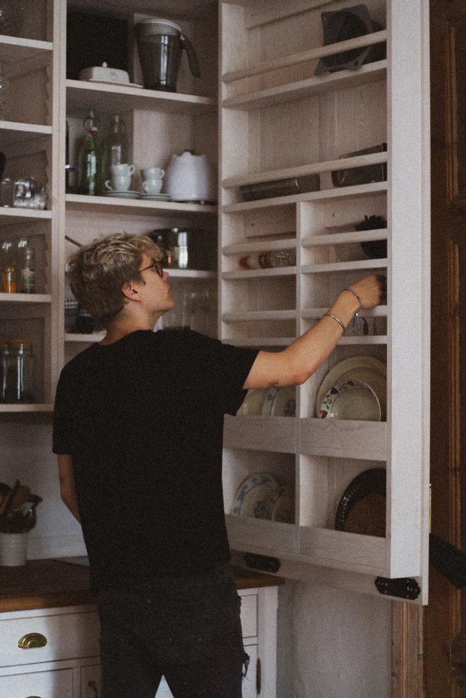 a blonde haired teen in black clothes with glasses standing infront of an open cabinet with pots, pans, dishes and cups