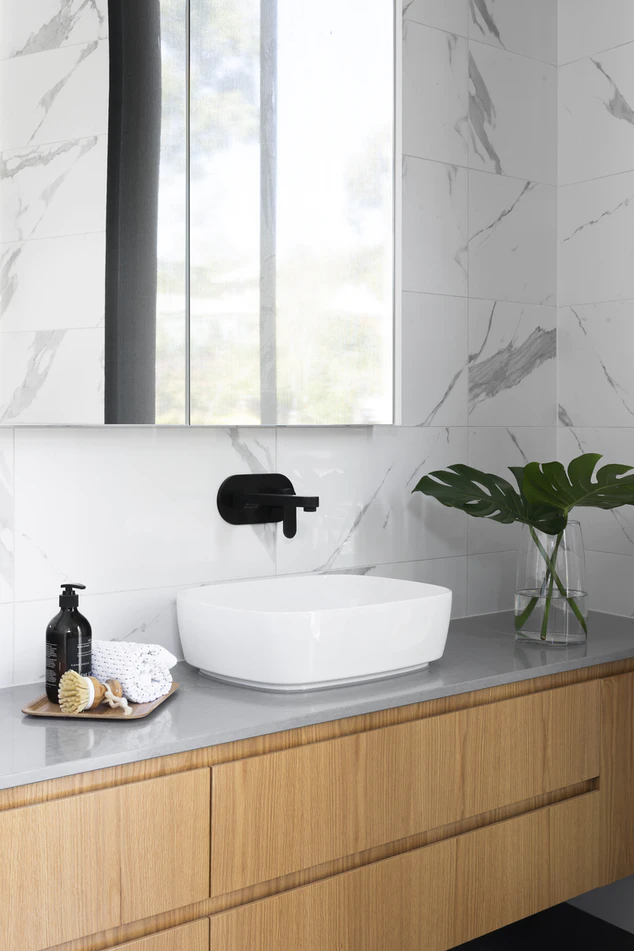 a bamboo bathroom benchtop with monstera leaves in a vase and a small white sink with a black tap