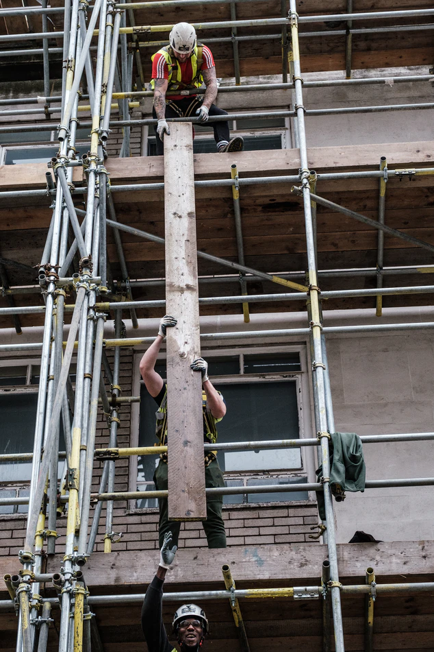 Three builders helping each other to lift a wooden plank up some scaffolding.